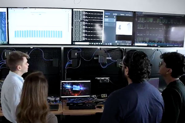 Four people stand in front of desks with computers and large monitors displaying data, graphs, and code in a technology lab setting.