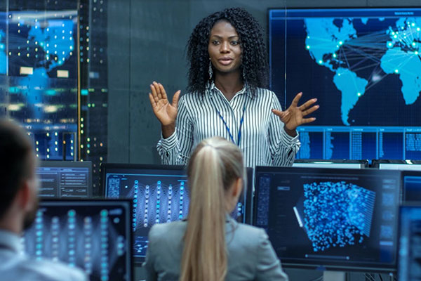 A woman stands confidently, gesturing while presenting in a control room filled with screens displaying data and a world map