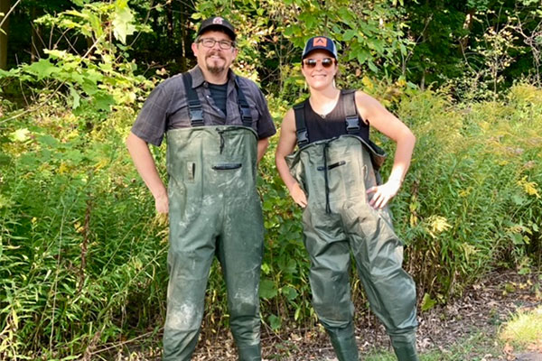 Two faculty members wearing green waders, standing in a forest