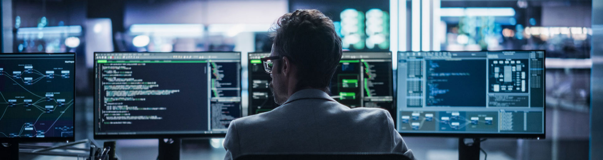 A person sits at a desk with multiple monitors displaying code in a dimly lit office