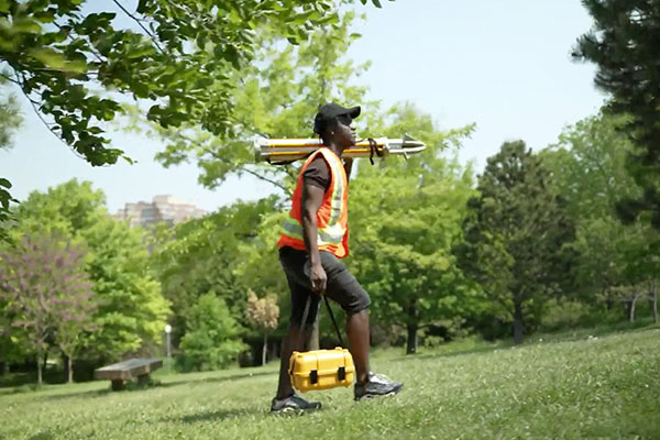 person in safety vest carrying surveying equipment through a grassy park
